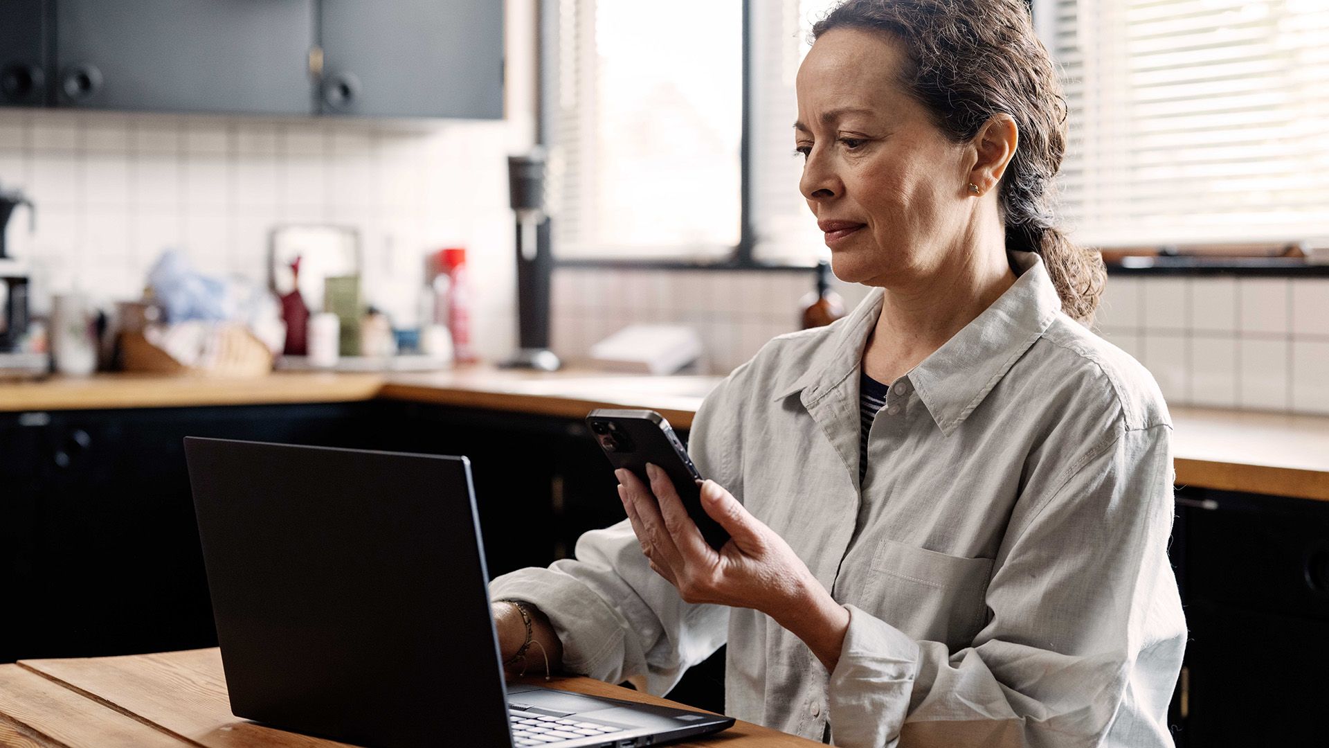Kvinde foran computer med telefon i hånden, kigger på cyberhjælp, der er en del af hendes indboforsikring